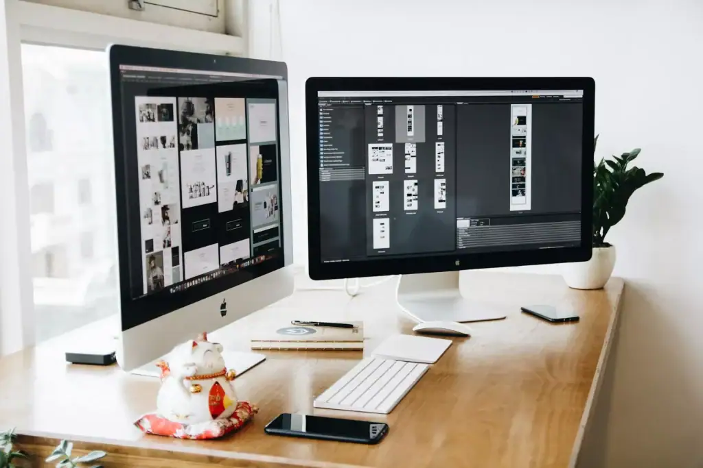 Desktop setup with two monitors displaying design projects, a keyboard, mouse, smartphone, and decorative cat figurine on a wooden desk.