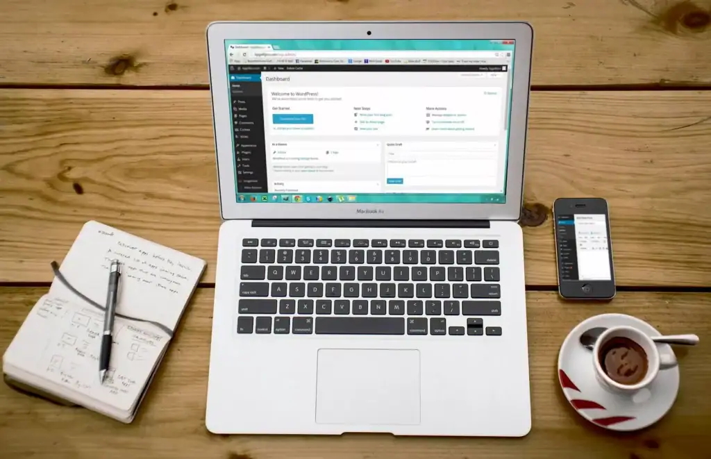 Laptop displaying WordPress admin dashboard, with a smartphone, notebook, and coffee on wooden table.
